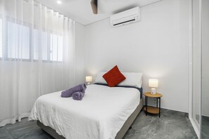 A hotel room with a bed, two bedside lamps, a red pillow, a rolled towel, and an air conditioning unit.