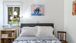 A bedroom with a bed, bedside lamps, a nightstand, a window with a view of trees, and a painting on the wall.