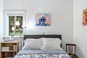 Un dormitorio con cama, lámparas de noche, mesita de noche, ventana con vista a árboles y un cuadro en la pared.