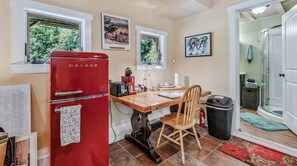 Cocina compacta con un refrigerador rojo Galanz, una mesa de comedor de madera y una silla. Hay ventanas con obras de arte encima, y una puerta que da a una zona de ducha.