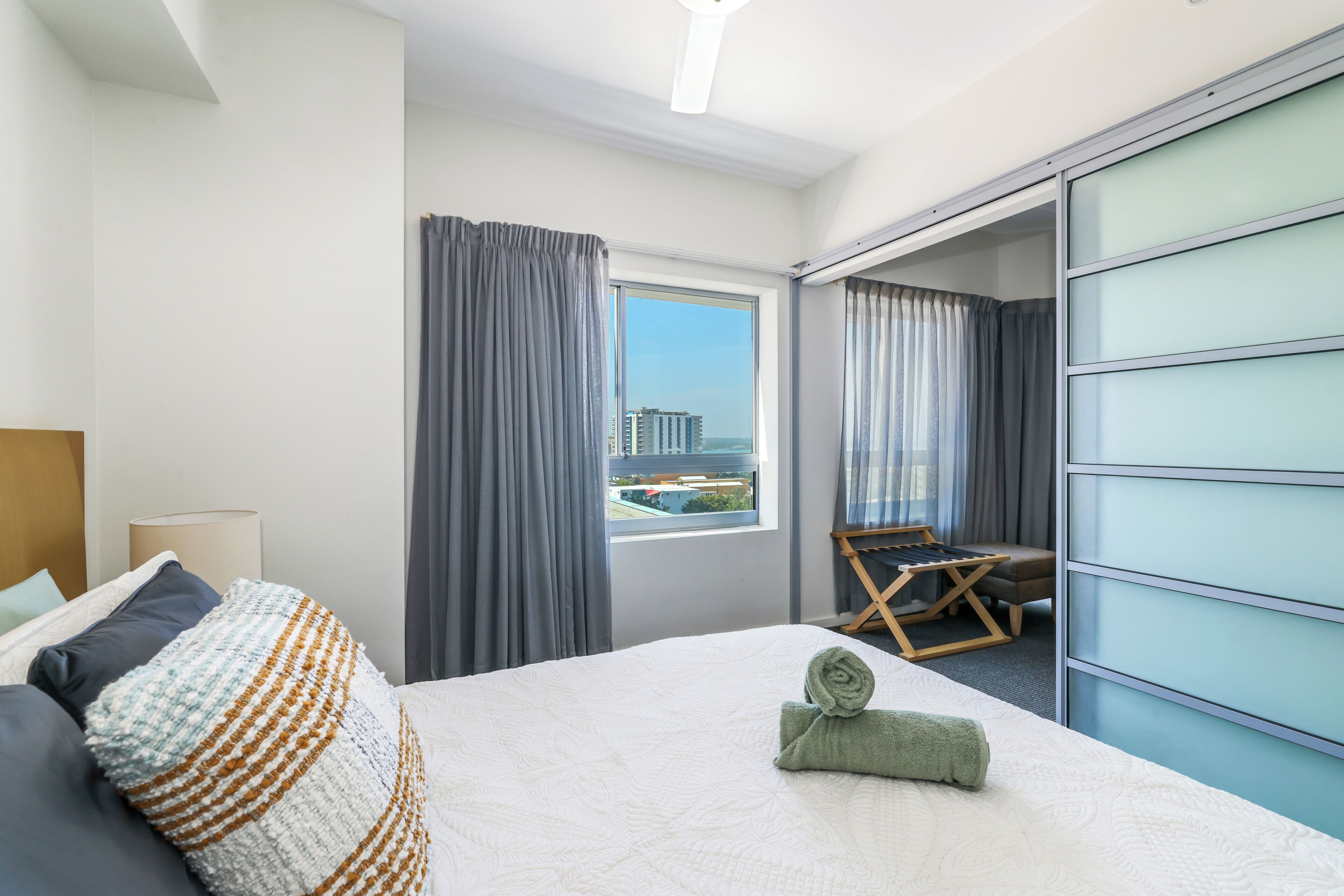 Ein Hotelzimmer mit Bett, Fenster mit Vorhängen, Blick auf Gebäude und einer Schiebetür zu einem weiteren Raum.