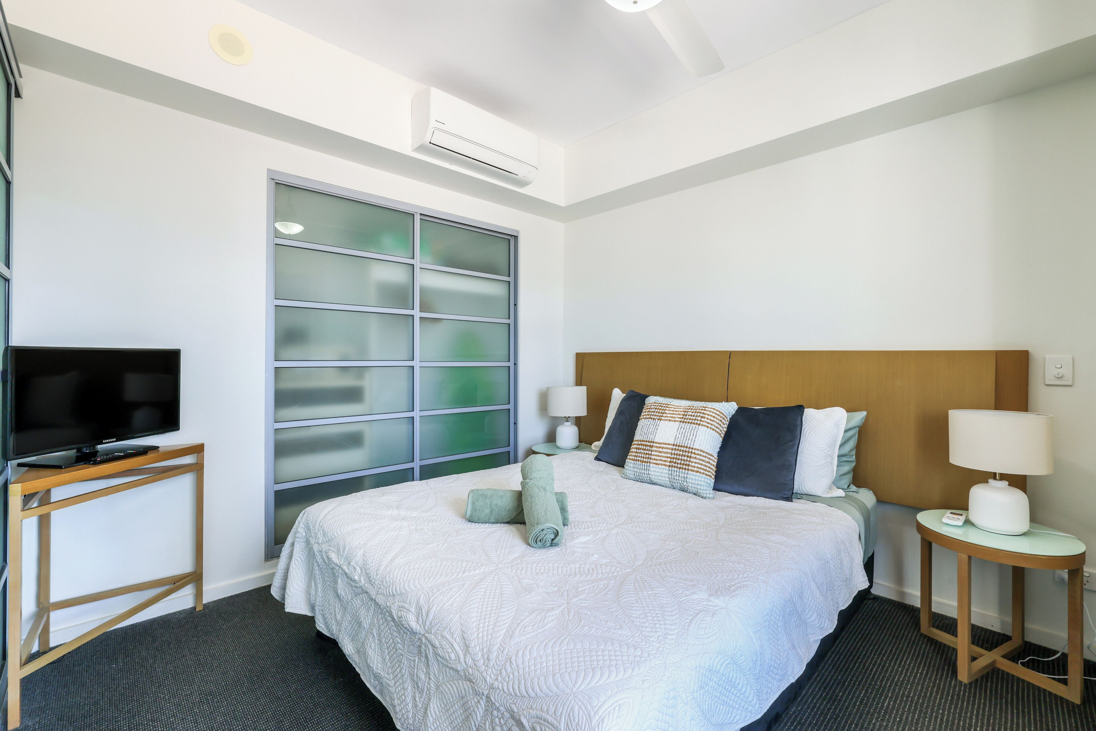 A hotel room with a bed, a television on a stand, a bedside table with a lamp, and a window with frosted glass panels.