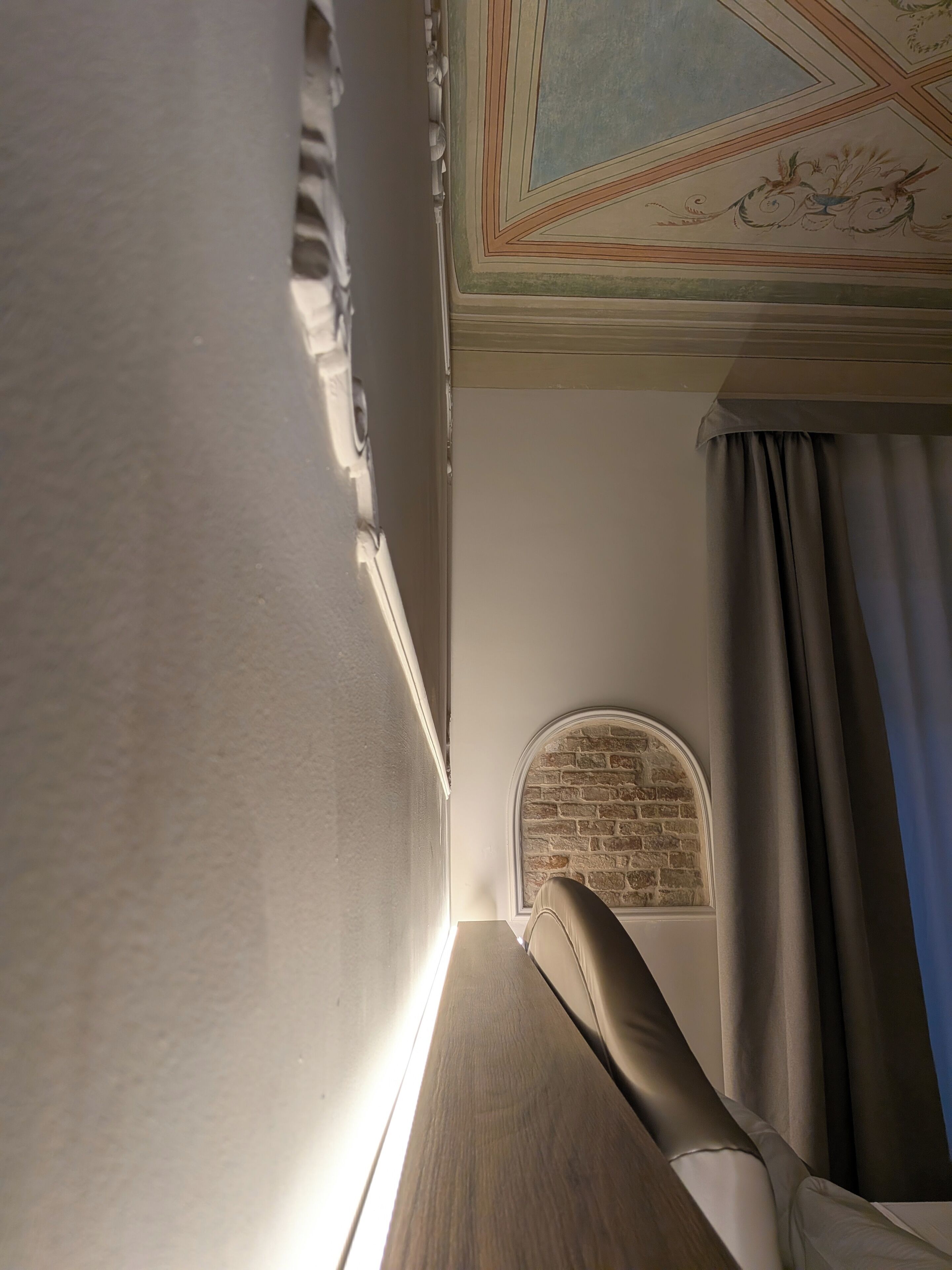 Ein Zimmer mit einem hölzernen Kopfteil, einem Fenster mit roher Ziegelwand und einer Decke mit ornamentalem Dekor.