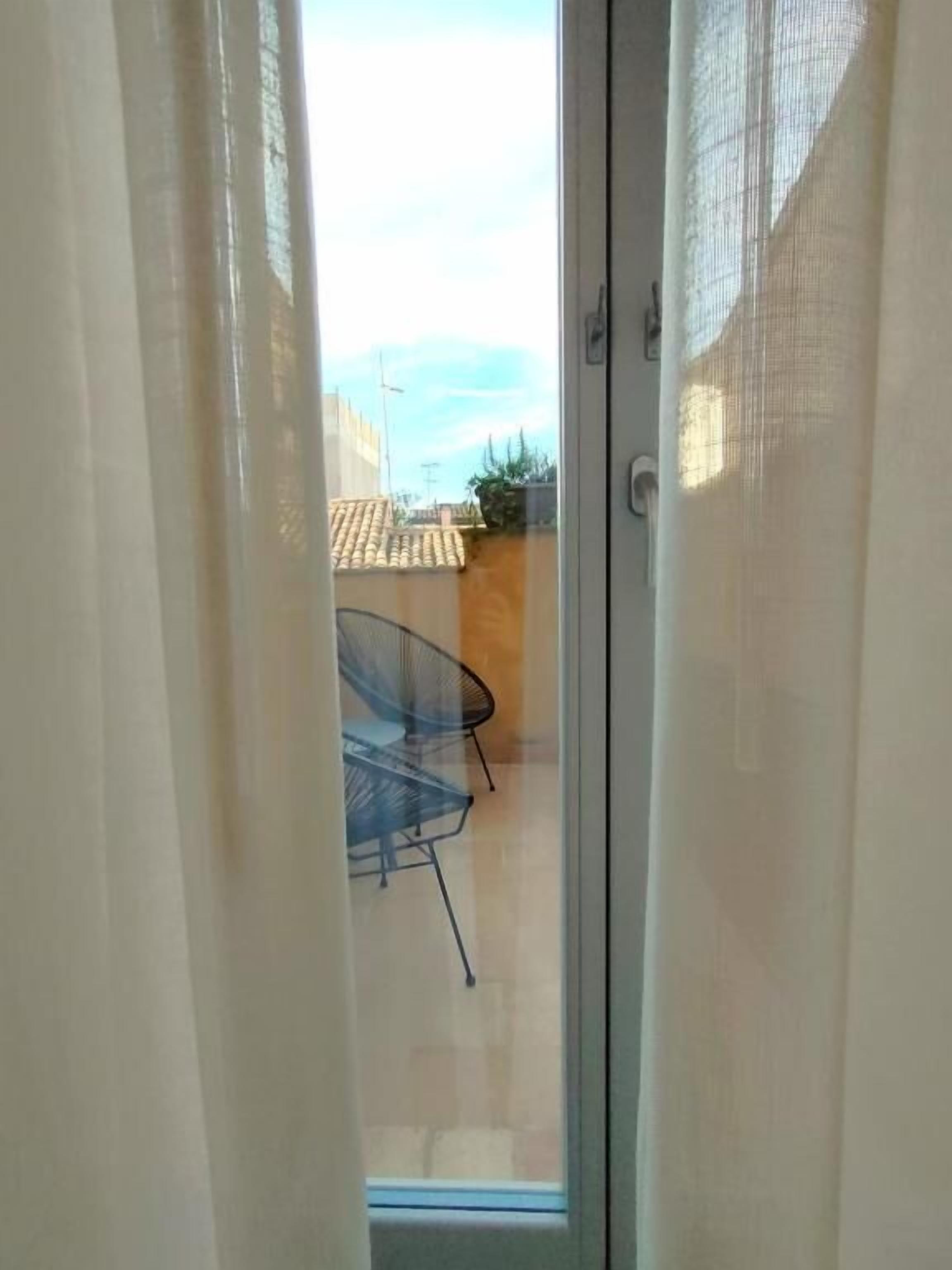 Vista a través de una ventana que muestra una puerta, una silla y un edificio con tejado de tejas al fondo.