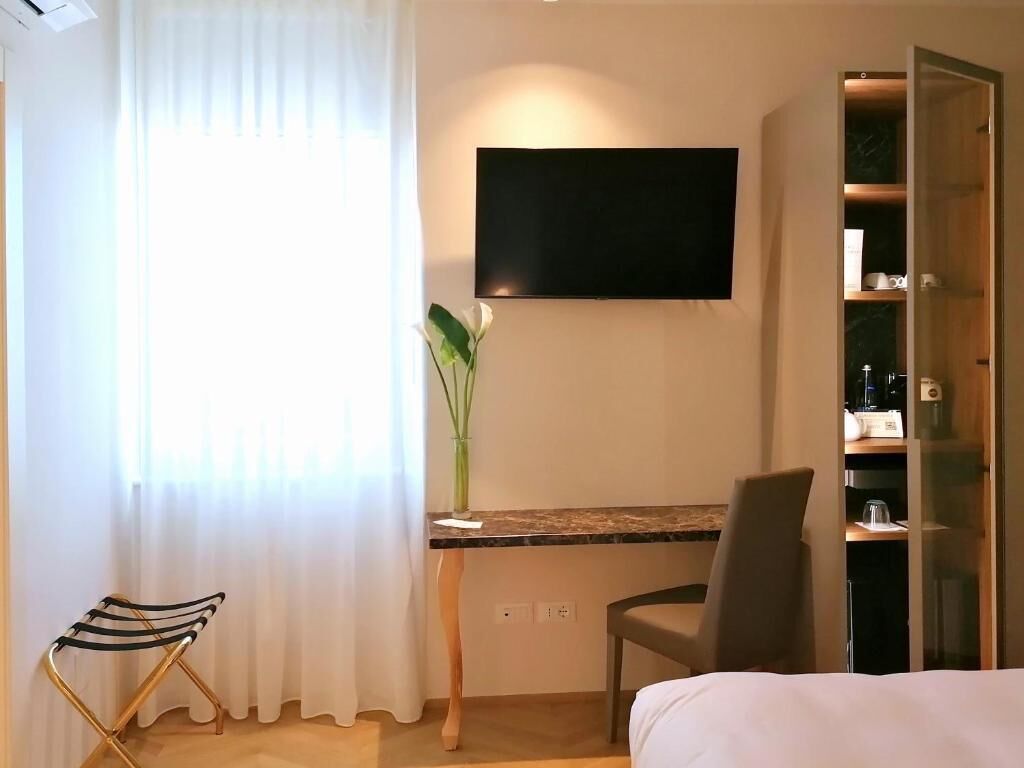 A hotel room with a bed, a desk with a chair, a television mounted on the wall, and a cabinet with shelves.