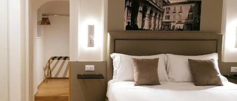 A hotel room with a bed, a nightstand, a wall-mounted picture, and a hallway leading to a staircase.