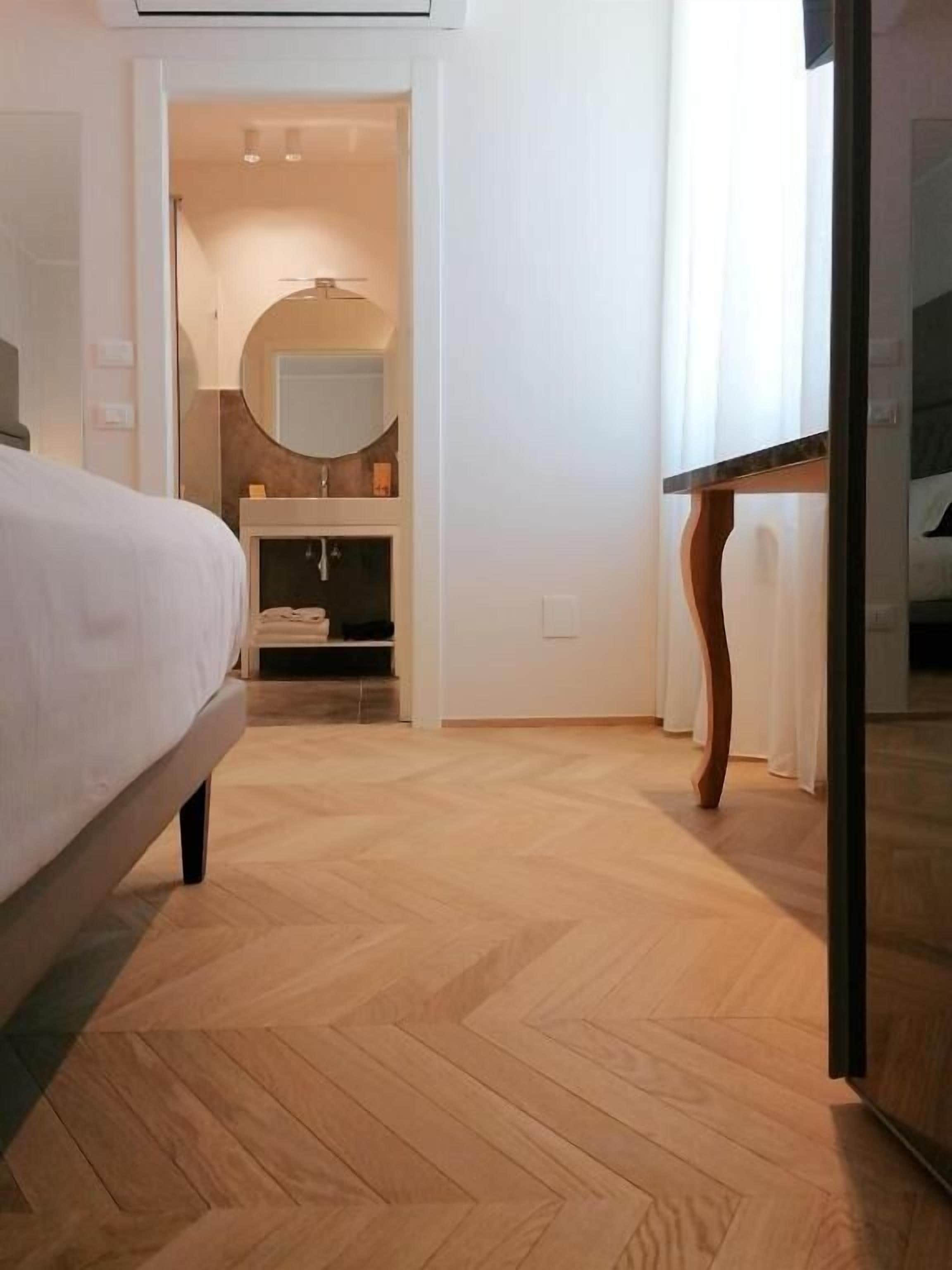 Een hotelkamer met een bed, een houten vloer, een badkamer met een spiegel en wastafel, en een deur naar een andere kamer.