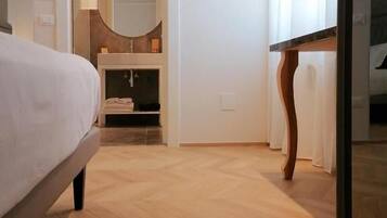 Een hotelkamer met een bed, een houten vloer, een badkamer met een spiegel en wastafel, en een deur naar een andere kamer.