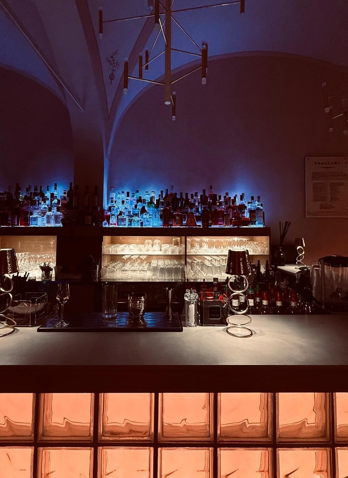Uma área de bar com prateleiras de garrafas de bebidas e copos, um balcão e um lustre.