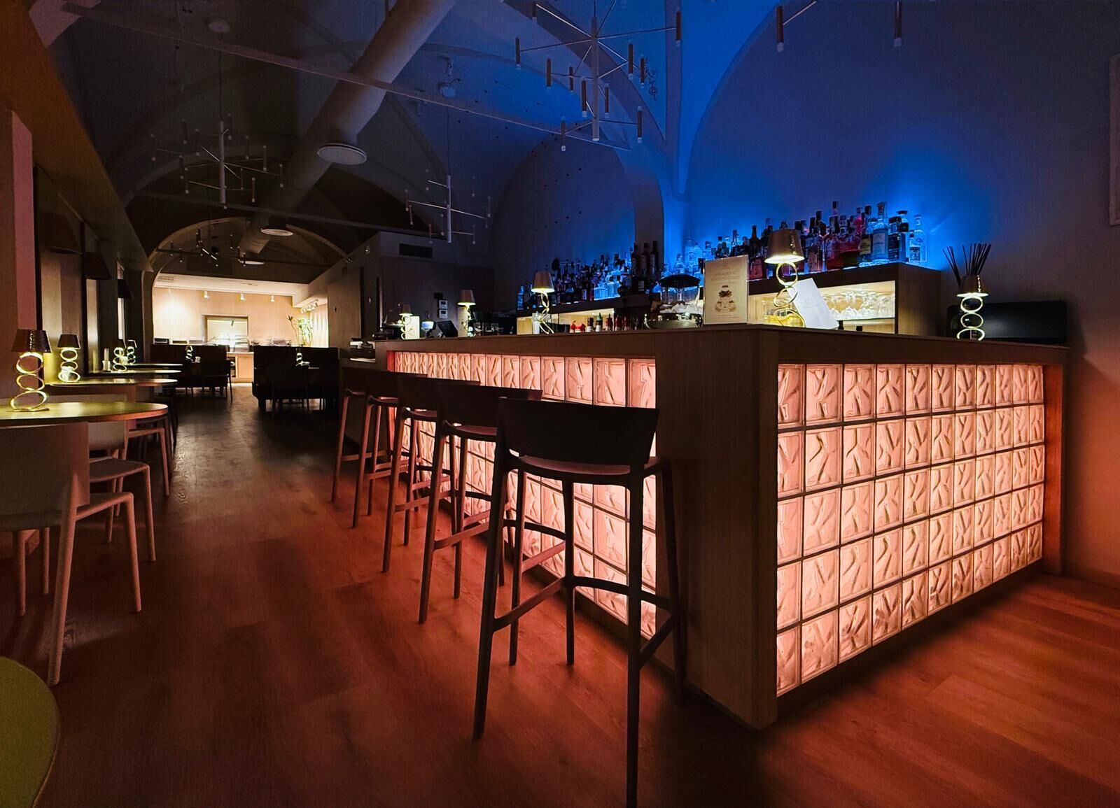 A bar area with wooden stools, a bar counter, and a variety of bottles.