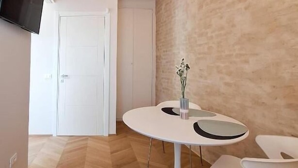 Una habitación con techo de madera, una mesa redonda blanca con sillas, un televisor de pantalla plana montado en la pared y una puerta que da a otra habitación.
