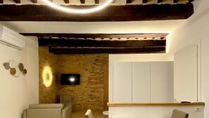Un comedor con una mesa y sillas blancas, un televisor montado en una pared de ladrillo y un techo con vigas de madera a la vista.