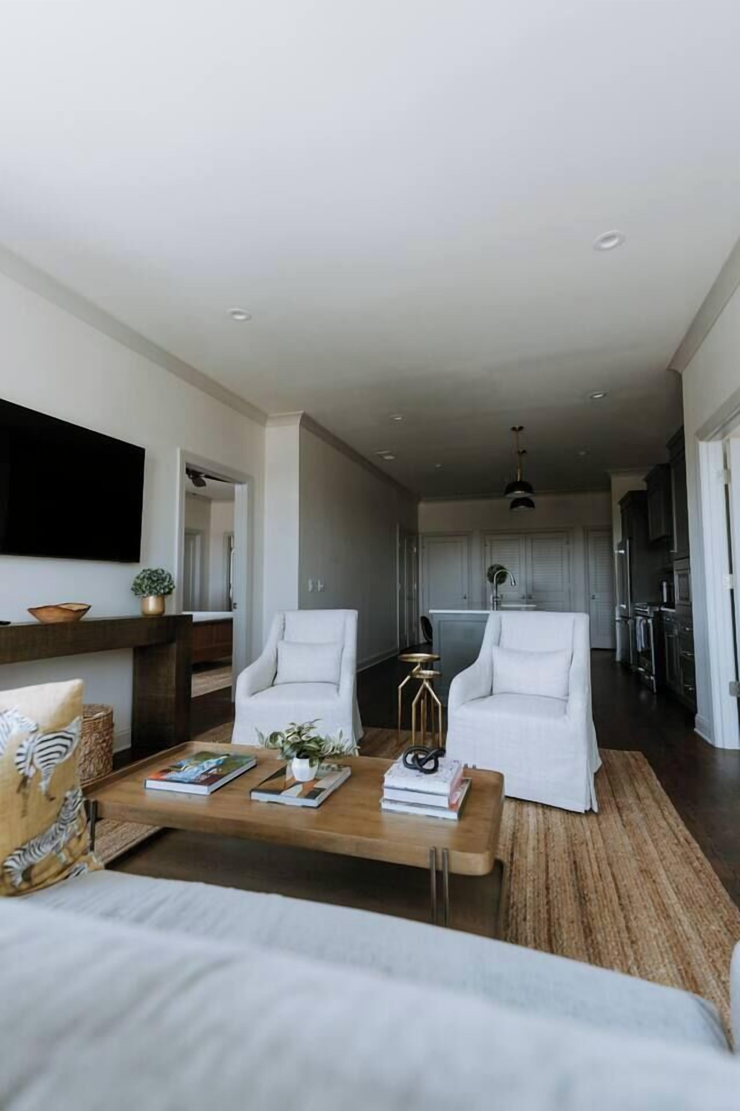 Een moderne woonkamer met een bank, twee witte fauteuils, een salontafel en een aan de muur bevestigde televisie.