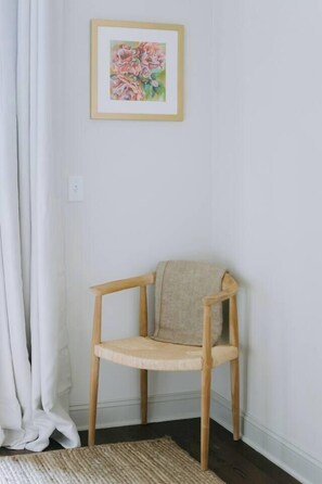 A corner room with a wooden chair, a framed floral artwork on the wall, and a white curtain.