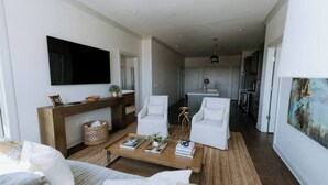 Uma sala de estar moderna com um sofá, mesa de centro e duas poltronas brancas. Uma televisão de tela plana está fixada na parede, e cozinha visível ao fundo.