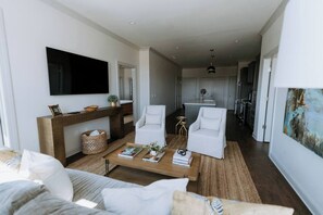 A modern living room with a sofa, coffee table, and two white armchairs. A flat-screen TV is mounted on the wall, and there is a kitchen visible in the background.