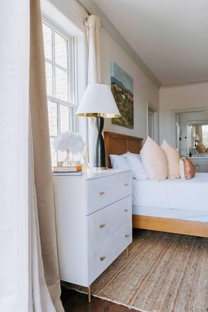 A bedroom with a bed, a bedside table with a lamp, a dresser, and a window with curtains.