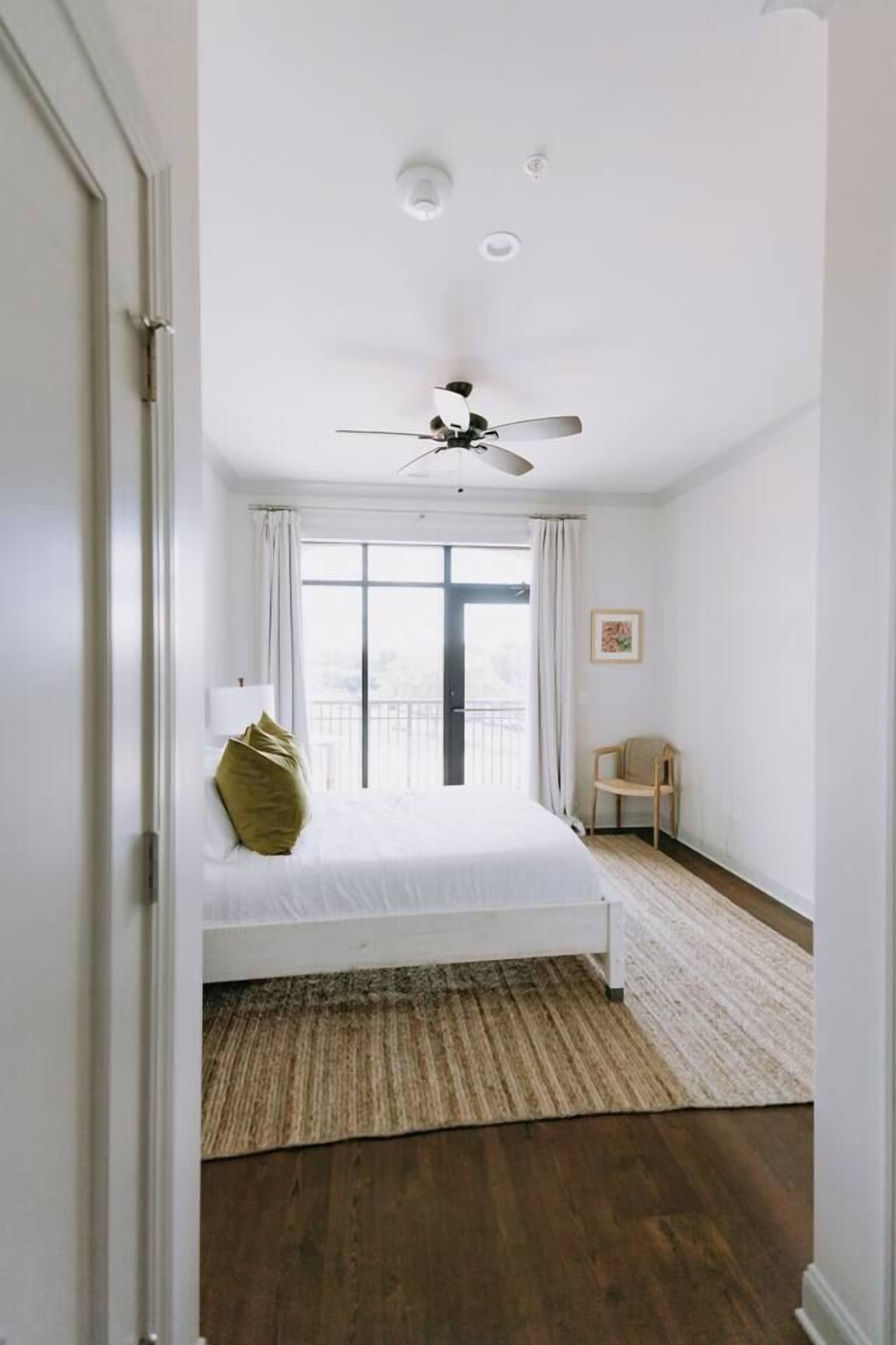 A bedroom with a bed, a ceiling fan, a window with curtains, a chair, and a door.