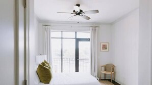 Quarto com cama, ventoinha no teto, janela com cortinas, uma cadeira e uma porta.