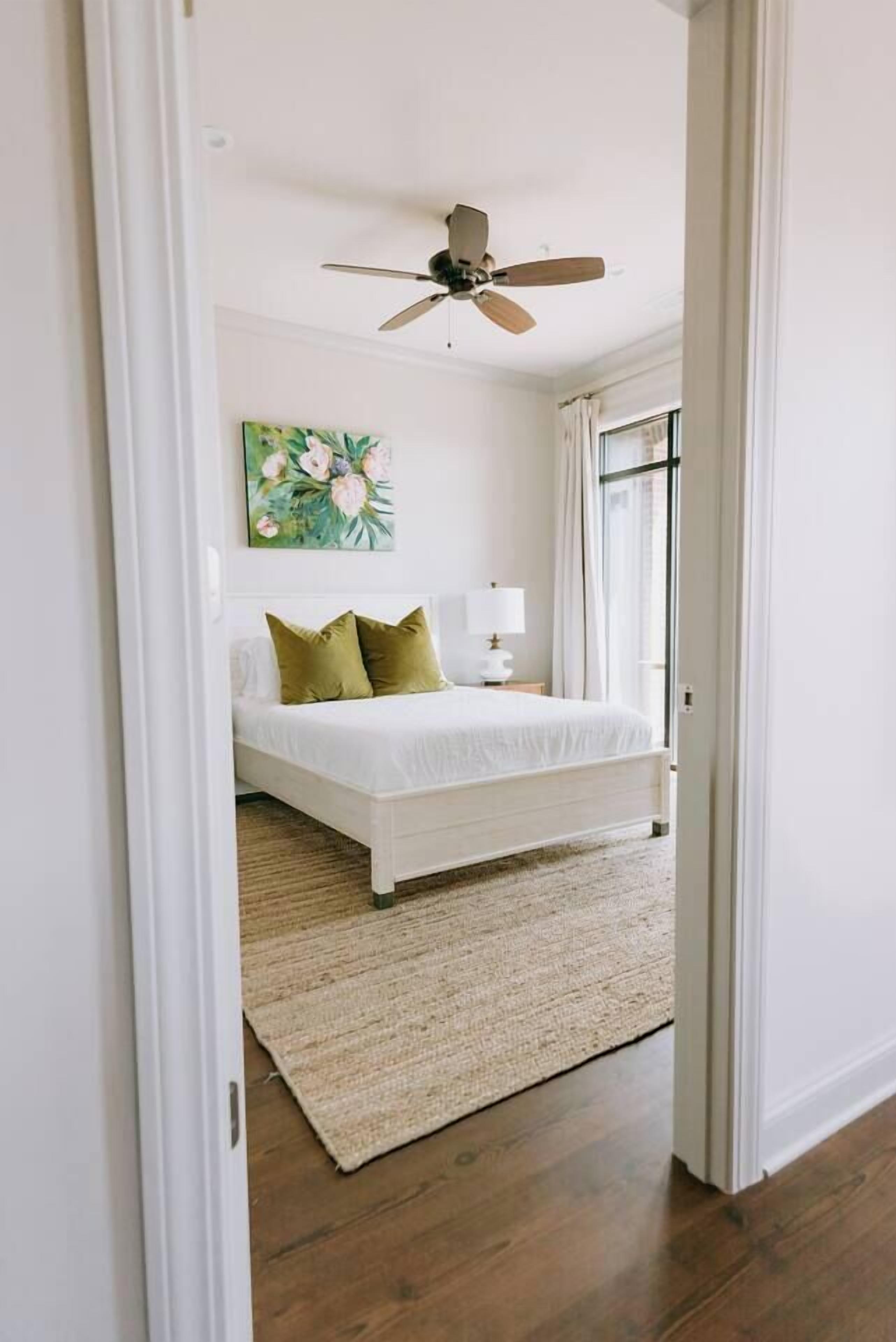 Un dormitorio con cama, ventilador de techo, un cuadro y una ventana con cortinas.