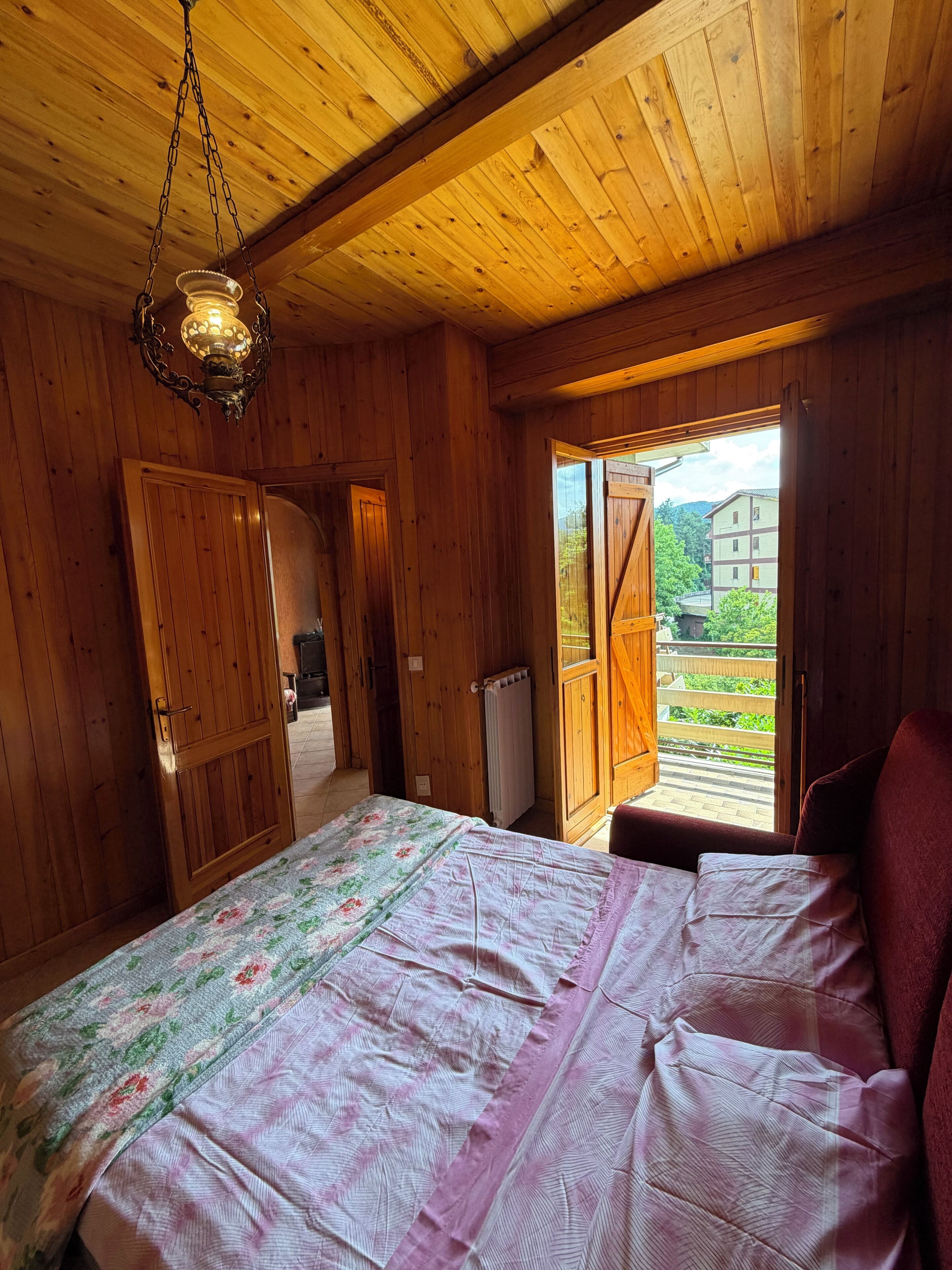 Habitació de fusta amb un llit, un lluminari penjant i vista cap a l'exterior.