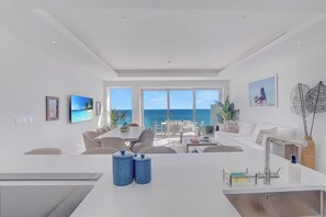 A modern living room with a large window overlooking the sea, a dining area with a view, and a kitchen with a sink and countertop.