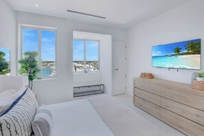 Ein modernes Schlafzimmer mit großem Fenster, einem Bett, einer hölzernen Kommode und einem Fernseher, der eine Strandansicht zeigt.