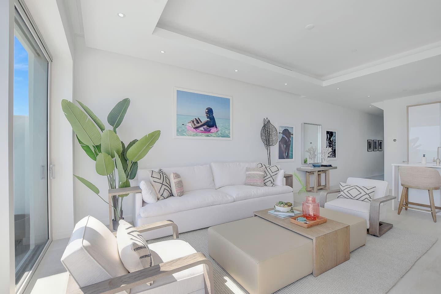 A modern living room with a white sofa, a coffee table, and a dining area with a chair. There is a large plant and artwork on the walls.