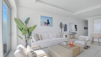 A modern living room with a white sofa, a coffee table, and a dining area with a chair. There is a large plant and artwork on the walls.