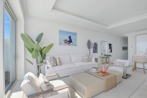 A modern living room with a white sofa, a coffee table, and a dining area with a chair. There is a large plant and artwork on the walls. - Palm Cay - 3BR Beachfront Penthouse + Private Pool (Nassau)