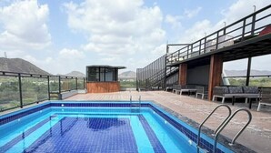 A rooftop swimming pool with a view of mountains, a wooden structure, and outdoor seating. - Tiger Hills Ridhira Retreats (Udaipur)