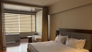 A hotel room with a large bed, a bedside table, a chair, a small table, and a window with blinds. - Tiger Hills Ridhira Retreats (Udaipur)