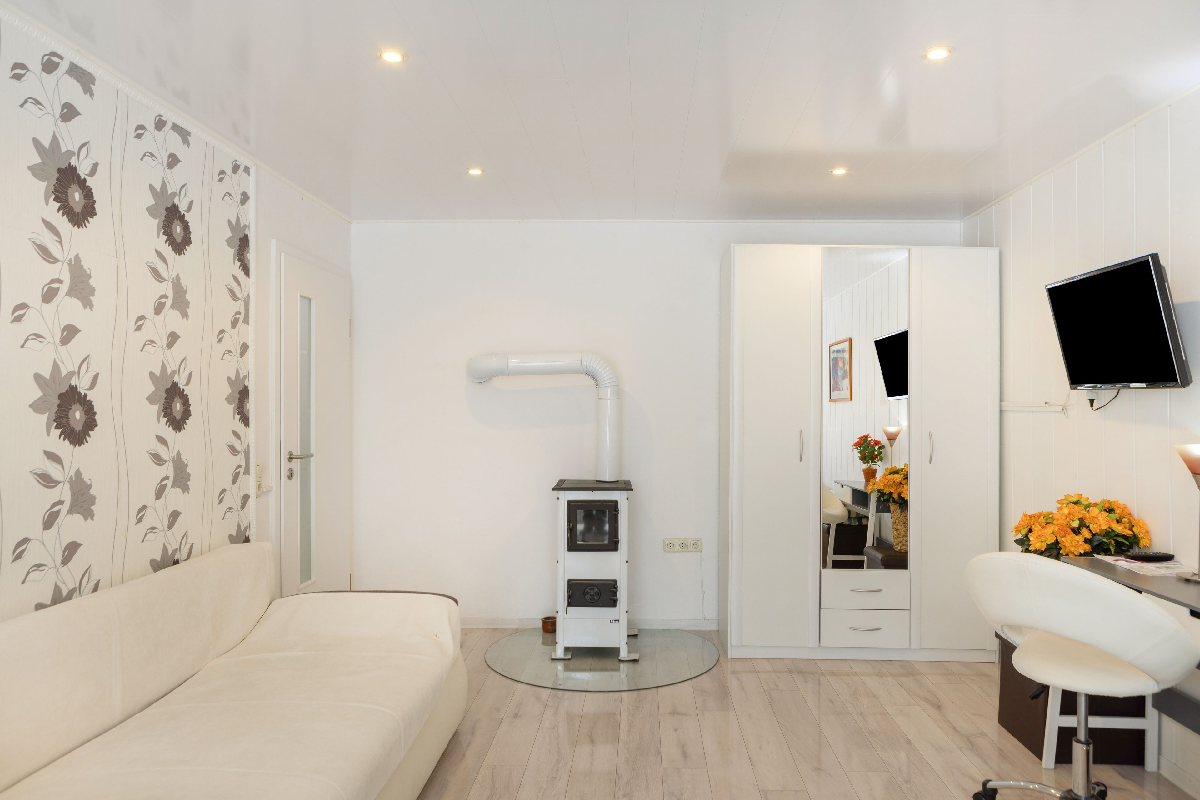 Una habitación moderna con un sofá blanco, un pequeño calentador, un televisor montado en la pared y un espejo con un jarrón de flores.