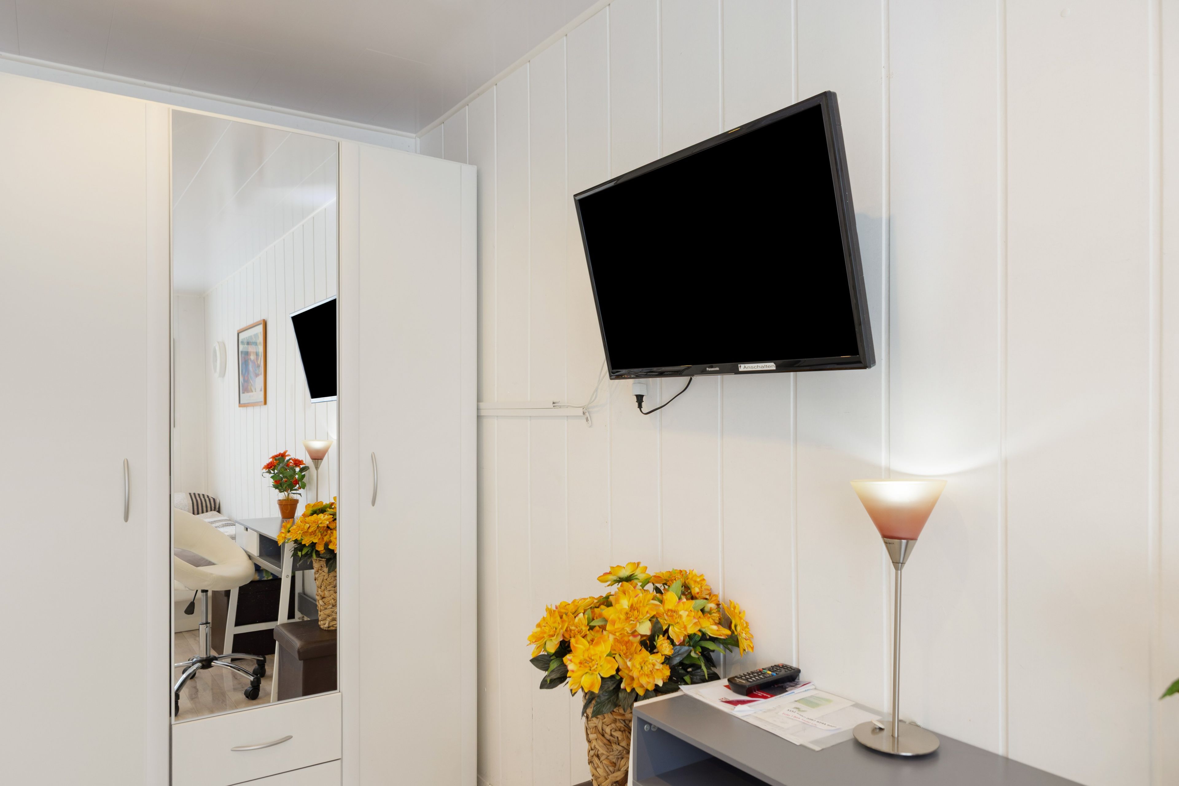 Ein Hotelzimmer mit einem an der Wand befestigten Flachbildfernseher, einem Schreibtisch mit Lampe und einem Spiegel, der den Raum reflektiert.