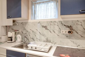 Eine Küche mit Marmor-Spritzschutz, einem fenster mit Vorhängen, einer Kaffeemaschine, einem Wasserkocher, einer Spüle und einem Geschirrständer.