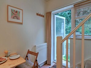 A room with a dining table, a chair, a radiator, a window with curtains, a wooden coat rack, and a staircase. - Apartment With Jacuzzi Bath (Torpoint)