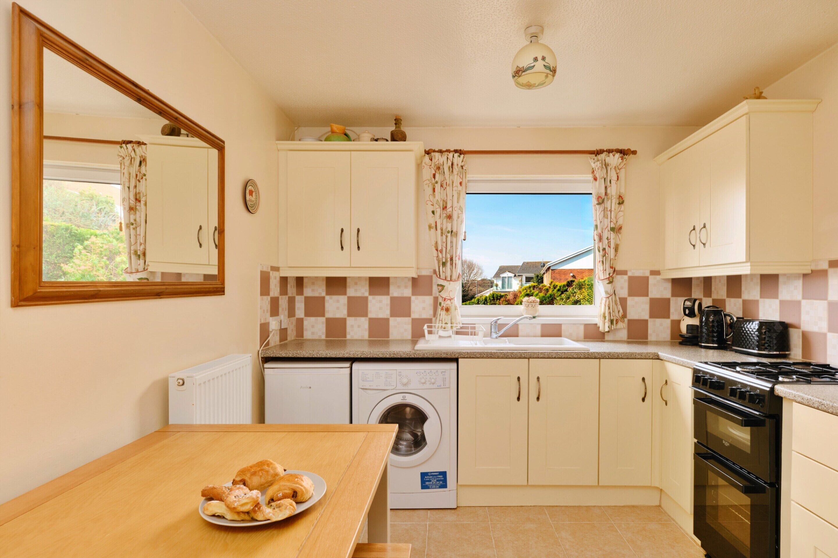A compact kitchen with a washing machine, a stove, and a dining area with a plate of pastries.