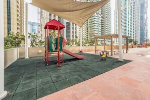 Un parque infantil al aire libre con un tobogán rojo y una estructura para escalar, rodeado de edificios altos.