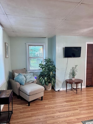 A living room with a sofa, a large potted plant, a television mounted on the wall, and a wooden floor. - Walk to Downtown & IU • Charming Retreat (Bloomington)