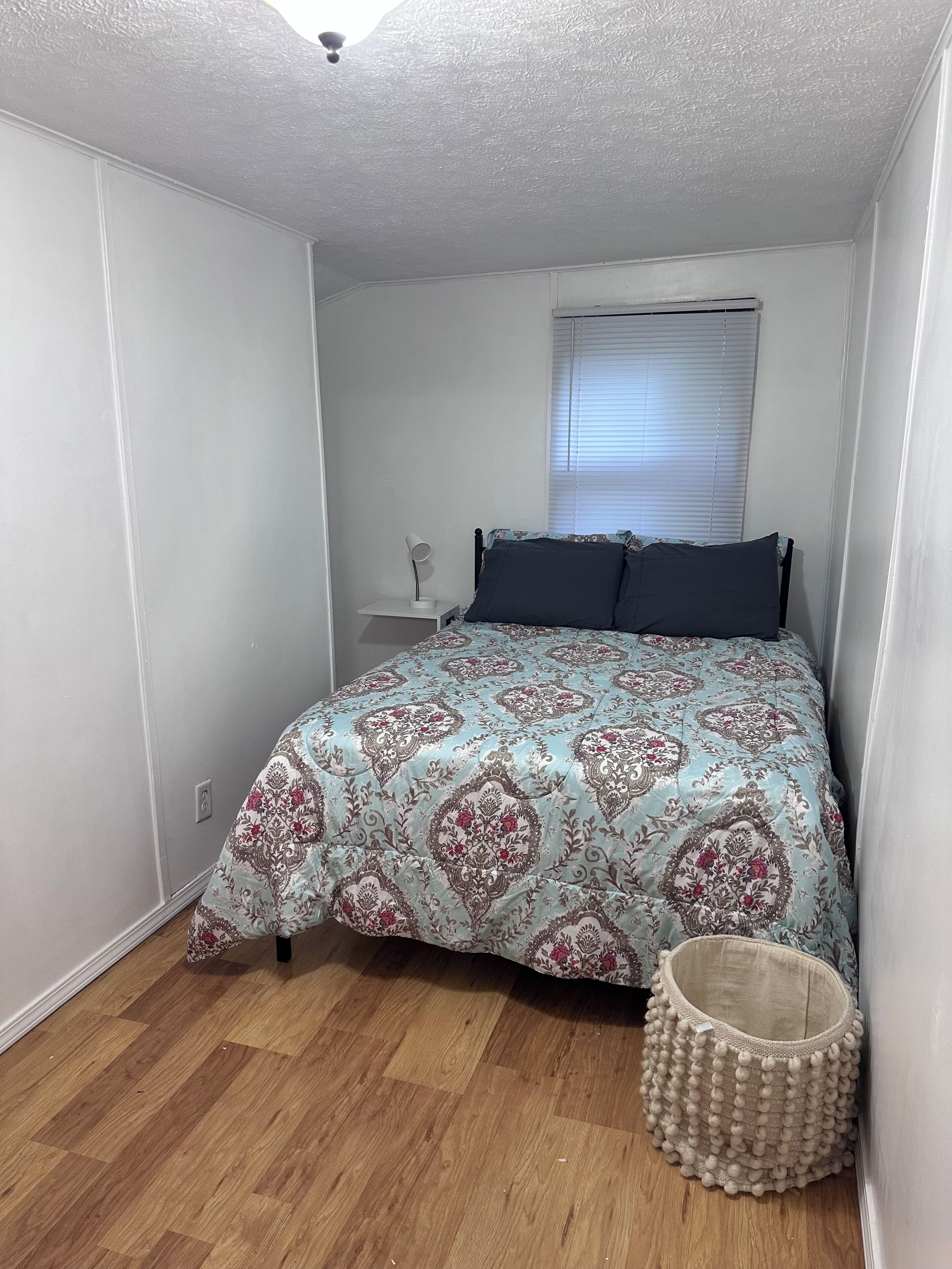 A bedroom with a bed, a bedside lamp, a window with blinds, a ceiling fan, and a small wooden floor.