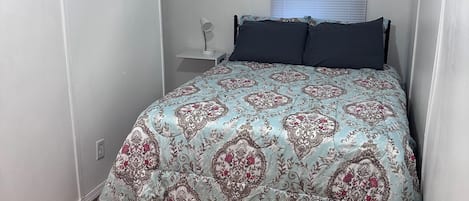 A bedroom with a bed, a bedside lamp, a window with blinds, a ceiling fan, and a small wooden floor.