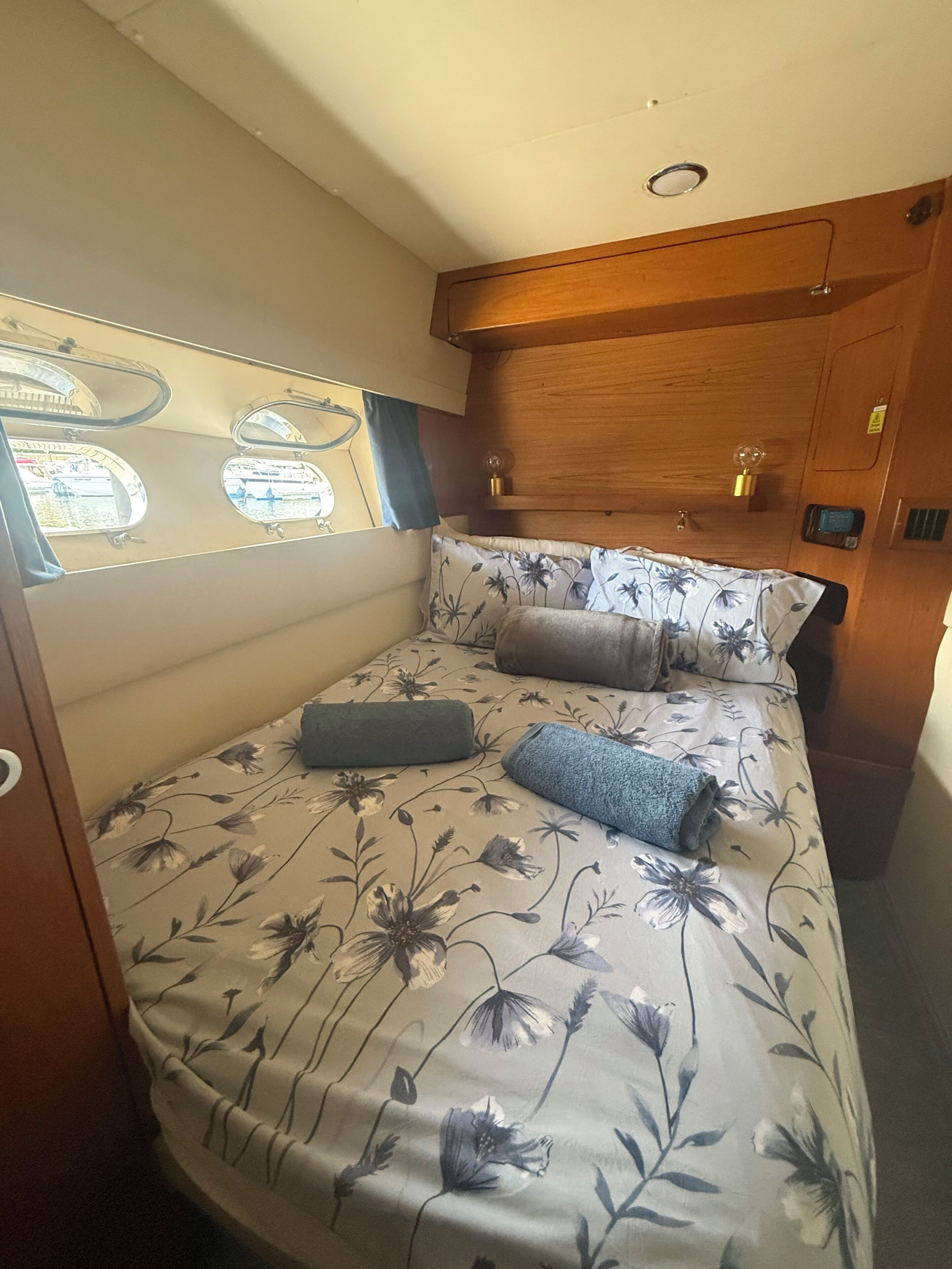 A compact, well-lit interior with a bed featuring floral bedding and two pillows, a small window with a view, and a wooden headboard.