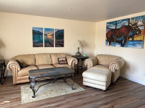 Ein Wohnzimmer mit einem beigefarbenen Sofa, einem passenden Sessel, einem Couchtisch und einem Teppich. An der Wand hängen drei gerahmte Bilder und ein einzelnes Gemälde.