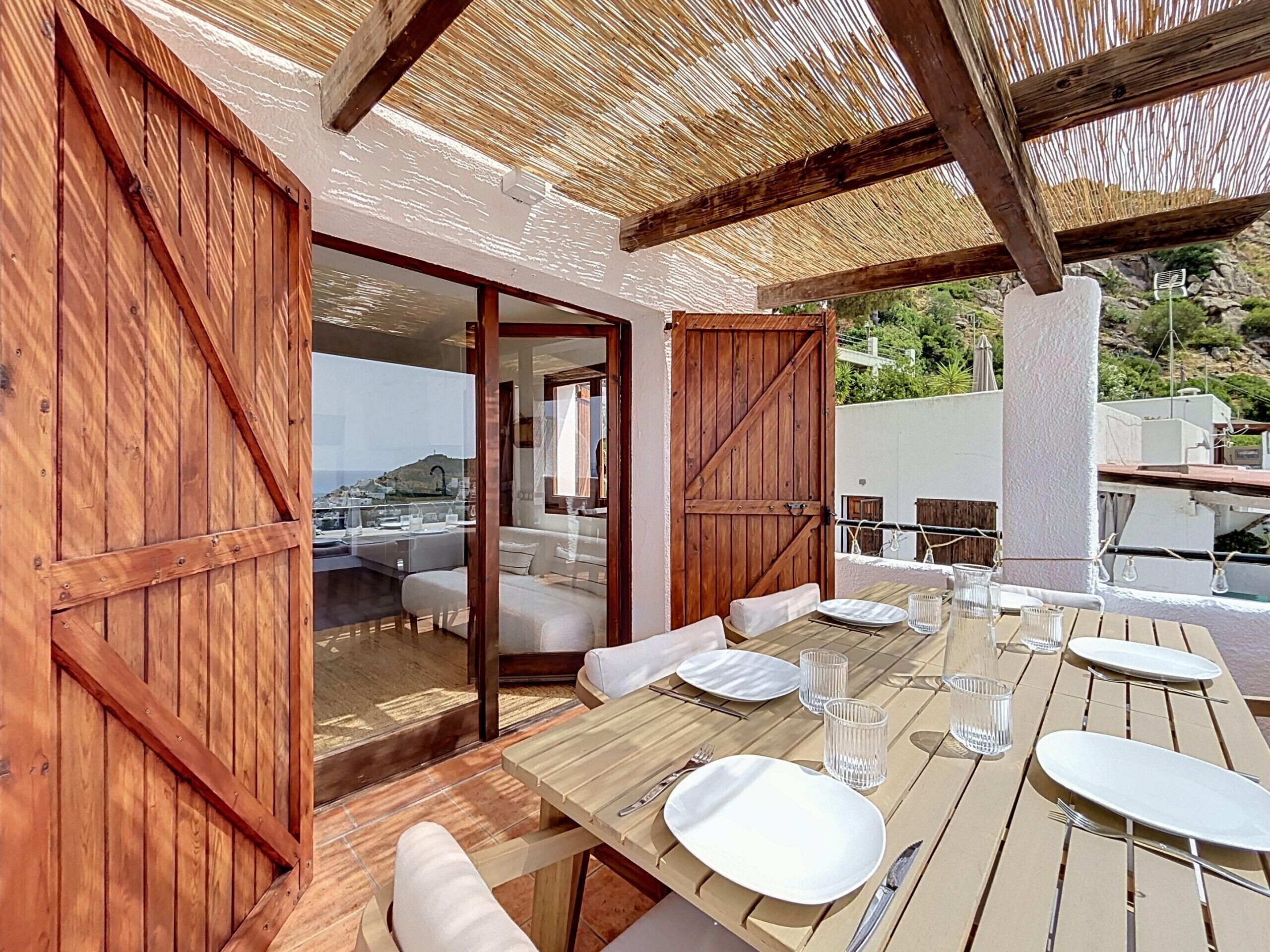 An outdoor dining area with wooden tables and chairs, a view of the sea, and a covered section with a wooden ceiling.