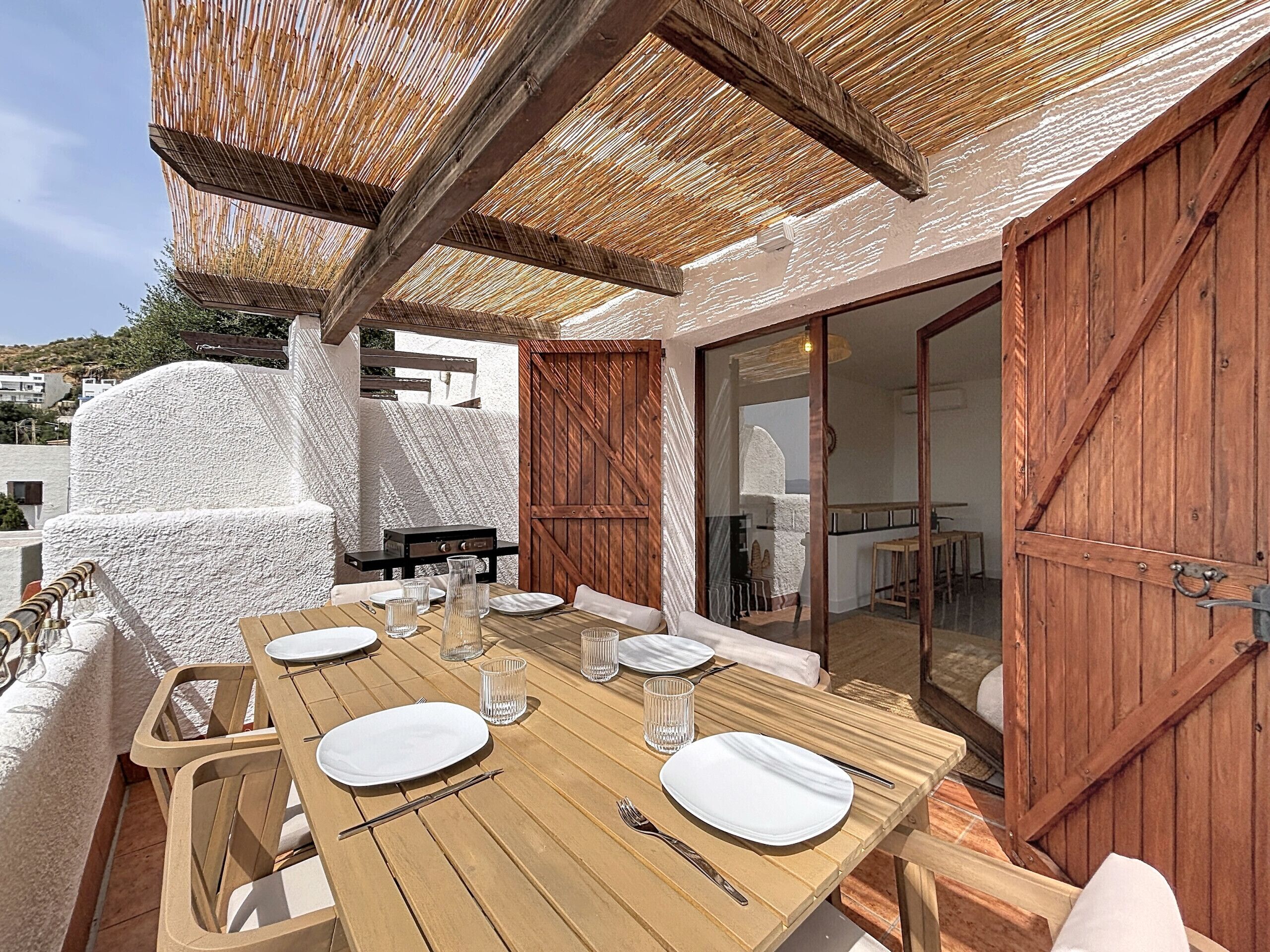 An outdoor dining area with a wooden table set for four, surrounded by wicker chairs, under a thatched roof.