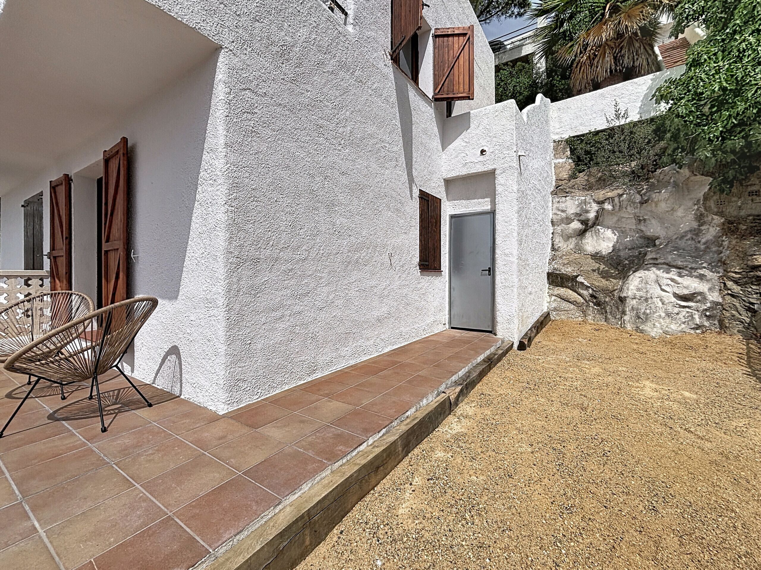 Un bâtiment blanc avec une porte et des fenêtres, une terrasse avec des chaises et une zone recouverte de gravier.