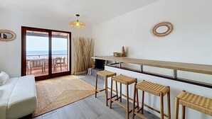 A modern living room with a sofa, a dining area with a high table and stools, a large window with a view, and a ceiling fan. - BRAMER 36 Rosas / Roses - INTERimmo (Rosas / Roses)