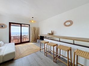 A modern living room with a sofa, a dining area with a high table and stools, a large window with a view, and a ceiling fan. - BRAMER 36 Rosas / Roses - INTERimmo (Rosas / Roses)