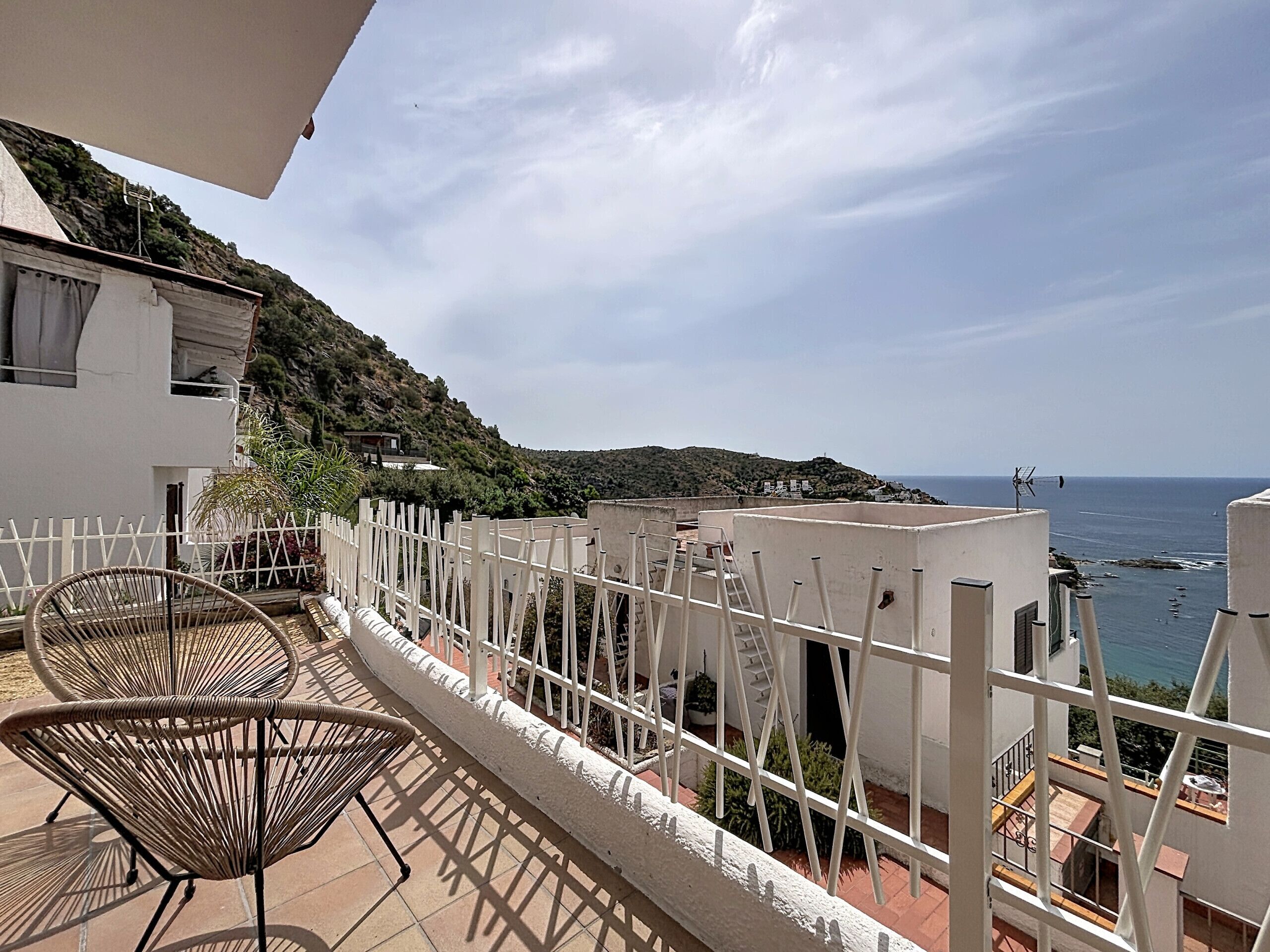 A balcony with a view of the sea, white railing, and two wicker chairs.
