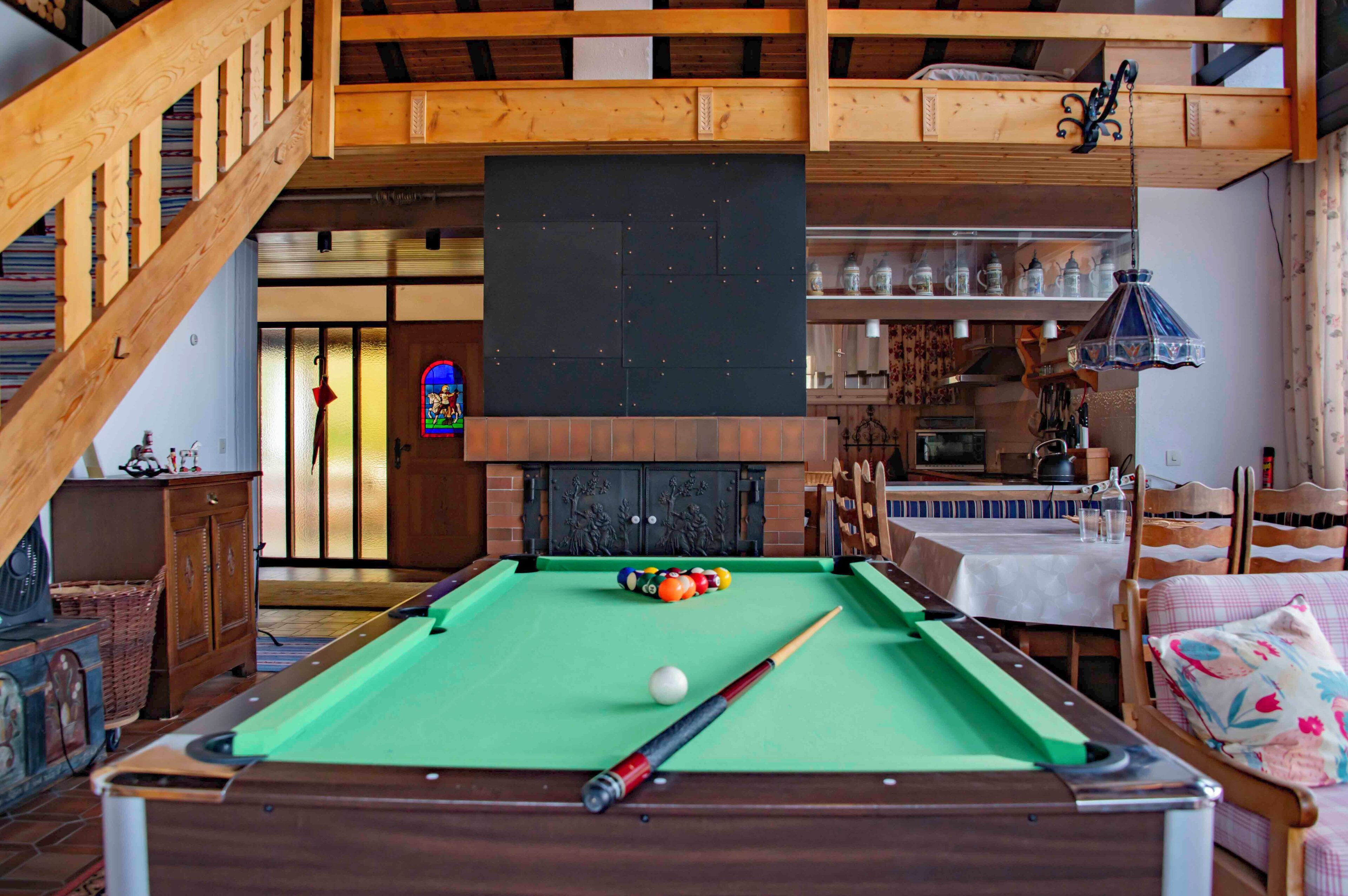 A pool table with balls, a billiard cue, and a fireplace in a room with wooden beams and a staircase.
